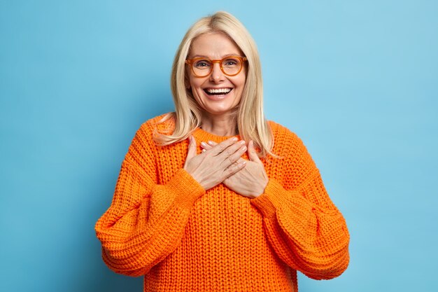 Portret van een blonde vrouw met vrolijke uitdrukking houdt de handen tegen de borst gedrukt en drukt zijn dankbaarheid uit voor het hartverwarmende compliment, draagt een bril en een oranje trui.