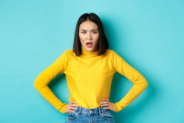 Portret van een boze en verwarde Aziatische vrouw die gefrustreerd naar de camera staart, staande tegen een blauwe achtergrond.