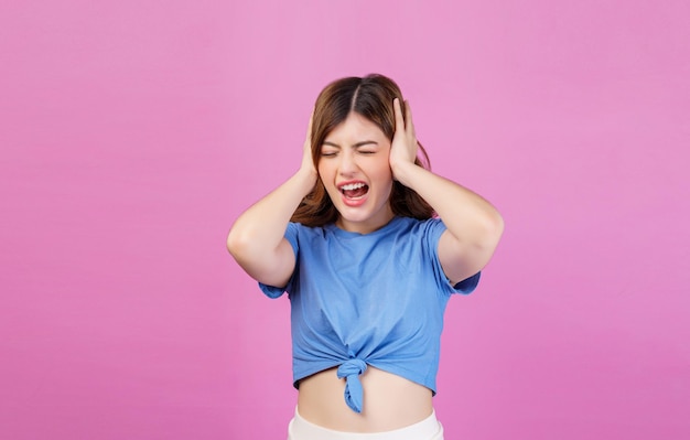 Portret van een boze geïrriteerde jonge vrouw die een casual t-shirt draagt dat haar oren bedekt met handen en schreeuwt terwijl ze geïsoleerd staat over roze achtergrond