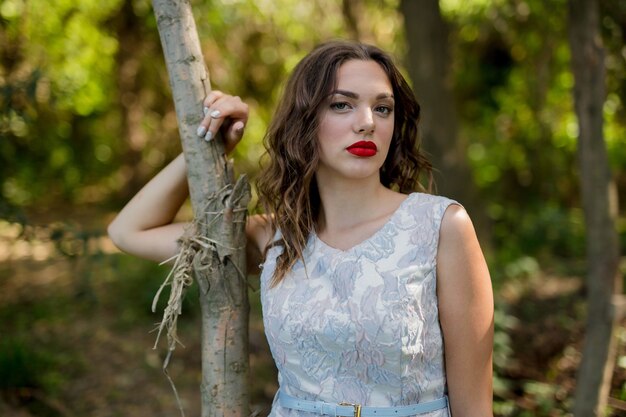 Portret van een brunette vrouw met een zomerjurk en rode lippenstift die een boomstam aanraakt