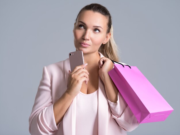 Portret van een denkende vrouw met roze boodschappentas en mobiele telefoon.