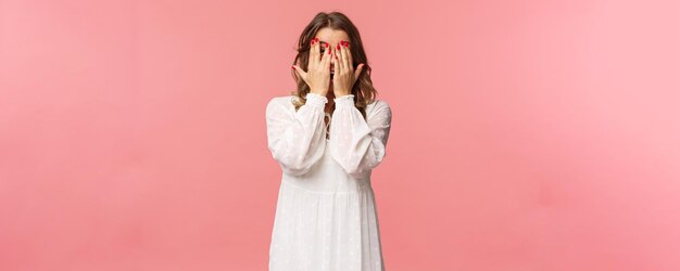 Portret van een dom schattig meisje met blond kort haar draagt een witte jurk die het gezicht achter de handen verbergt en belooft te wachten op een signaal, maar door de vingers gluurt, kan de verleiding niet weerstaan en staat roze achtergrond