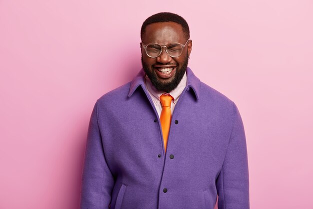 Portret van een donkere mannelijke werknemer heeft hysterische lach, positieve stemming, glimlacht breed, draagt paarse jas, oranje stropdas