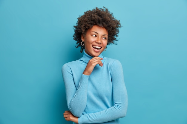 Portret van een donkere vrolijke vrouw met krullend haar, raakt kin zachtjes, lacht gelukkig, geniet van vrije dag, voelt zich gelukkig en enthousiast, hoort iets positiefs, draagt casual blauwe coltrui