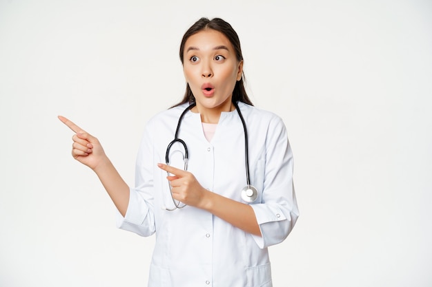 Portret van een enthousiaste vrouwelijke arts, een aziatische arts die naar links wijst met een verbaasde, verbaasde gezichtsuitdrukking, staande in een medisch gewaad tegen een witte achtergrond.