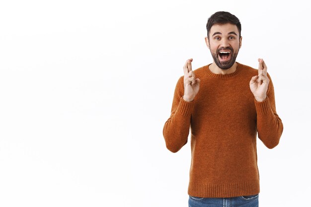 Portret van een enthousiaste, wensvolle en gelukkige jonge blanke man met baard, anticiperend op smaak en goed nieuws, investeren in iets hoop geld brengen, vingers kruisen en wachten