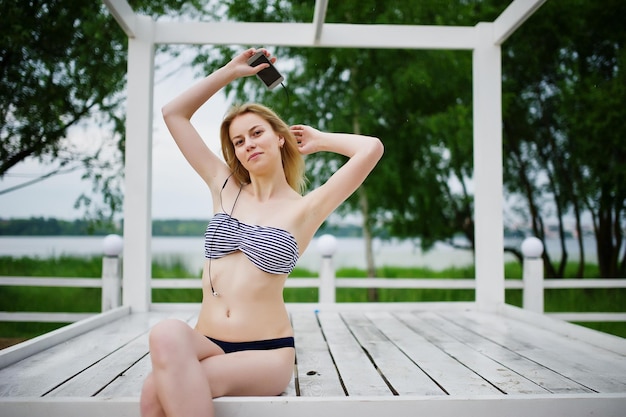 Gratis foto portret van een fantastische jonge vrouw in bikini zittend en poserend op een wit houten prieel in het park naast het meer