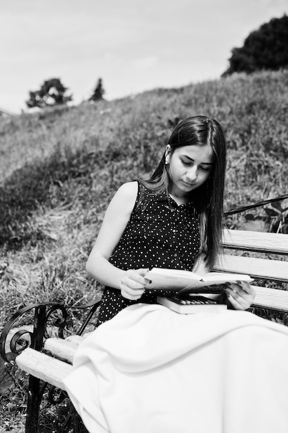 Gratis foto portret van een fantastische vrouw in zwarte polka dot jurk zittend op de bank met een enorme witte sjaal en lezen in het park zwart-wit foto