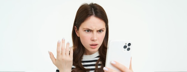 Gratis foto portret van een geïrriteerd meisje dat naar het scherm van haar mobiele telefoon staart met een gefrustreerd gezicht op een witte achtergrond