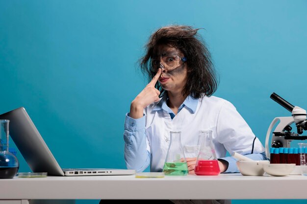 Portret van een gekke, grappige chemicus die grappig doet en een vies gezicht en rommelig haar heeft terwijl hij aan het bureau zit en naar de camera kijkt. Gekke wetenschapper met wild uiterlijk doet gek na explosie in laboratorium