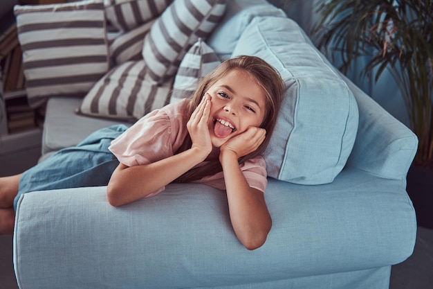 Portret van een gelukkig klein meisje met lang bruin haar en doordringende blik, toont tong op de camera, liggend op een bank thuis.