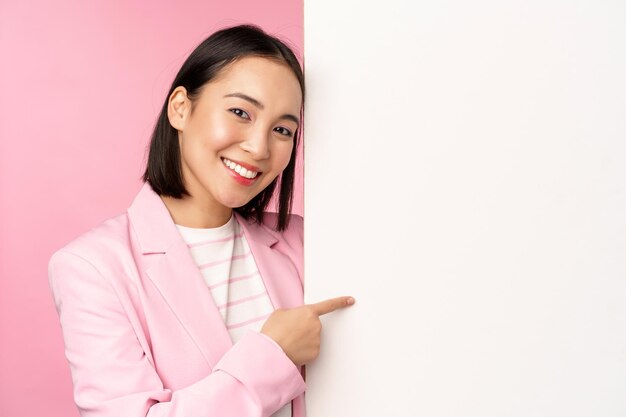 Portret van een gelukkig lachende Japanse zakelijke vrouw die met de vinger naar het bord wijst en iets laat zien op lege ruimte in pak over roze achtergrond