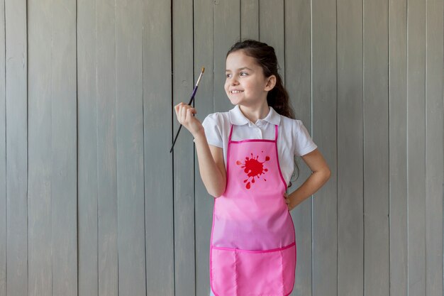 Portret van een gelukkig meisje in roze schort van de de holdingsverf van de schortholding in hand weg kijkend