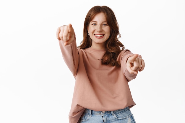 Portret van een gelukkig roodharig meisje dat jou kiest, met de vingers naar de camera wijst en glimlacht, uitnodigend voor een evenement, wenken om mee te doen, mensen rekruteren, we willen dat je gebaar maakt, staande op een witte achtergrond