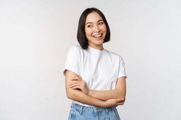 Portret van een gelukkige aziatische vrouw die lacht en zelfverzekerde kruisarmen op de borst stelt die tegen de achtergrond van de studio staan