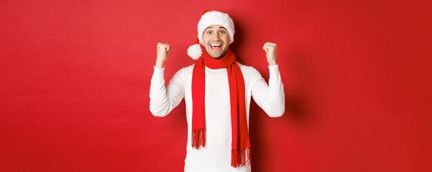Portret van een gelukkige en opgewonden man in kerstmuts en sjaal, zich verheugend en iets winnend, nieuwjaar vierend, staande op rode achtergrond