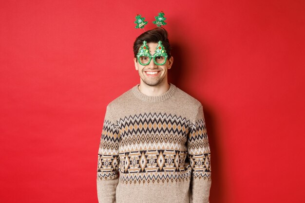 Portret van een gelukkige en schattige man in een wintertrui en een feestbril, nieuwjaar of kerstmis vierend, glimlachend terwijl hij over een rode achtergrond staat