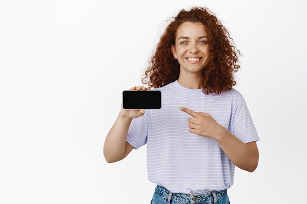 Portret van een gelukkige en trotse roodharige vrouw die met de vinger wijst naar het scherm van de mobiele telefoon, horizontaal smartphonedisplay, met een winkel- of applicatie-interface op wit.