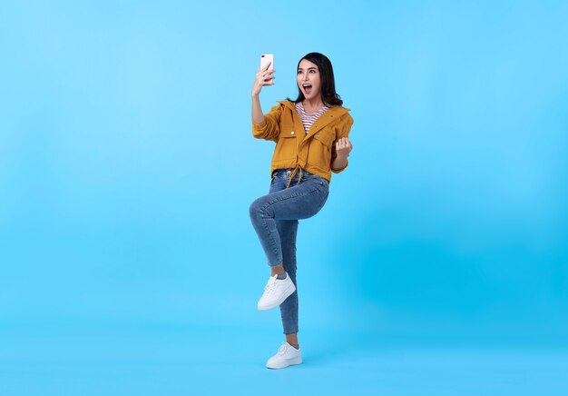 Portret van een gelukkige jonge Aziatische vrouw die met mobiele telefoon viert die over blauwe achtergrond wordt geïsoleerd.