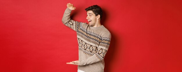 Portret van een gelukkige jonge man in een kersttrui, met een groot cadeau, glimlachend en verbaasd over een cool cadeau, staande op een rode achtergrond.