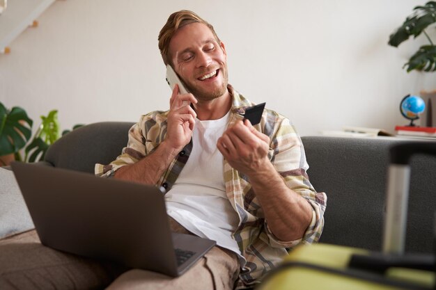 Portret van een gelukkige jonge toerist met een laptop en een creditcard die aan het bellen is op een mobiele telefoon