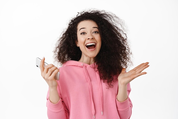 Portret van een gelukkige jonge vrouw die smartphone gebruikt, zich verheugt en schreeuwt van vreugde, geld wint op mobiele telefoon, zegeviert, wint op smartphone, staande tegen een witte achtergrond