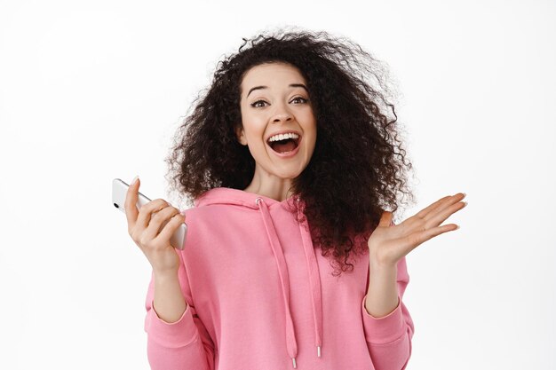 Portret van een gelukkige jonge vrouw die smartphone gebruikt, zich verheugt en schreeuwt van vreugde, geld wint op mobiele telefoon, zegeviert, wint op smartphone, staande tegen een witte achtergrond