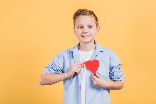 Portret van een gelukkige jongen die rood hart toont dichtbij zijn borst tegen gele achtergrond