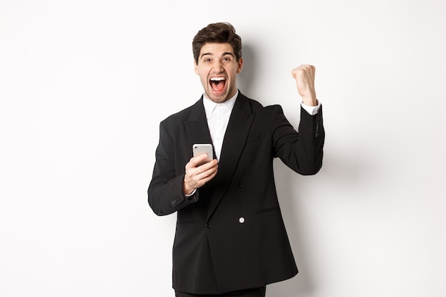 Portret van een gelukkige knappe man in pak, vreugde, doel bereiken op mobiele app, vuist opheffen en ja schreeuwen, smartphone vasthouden, staande tegen een witte achtergrond
