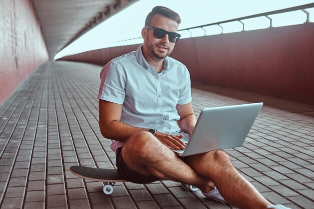 Portret van een gelukkige knappe modieuze freelancer in zonnebril gekleed in een wit overhemd en korte broek die op een laptop werkt terwijl hij op een skateboard onder de brug zit, kijkend naar een camera.