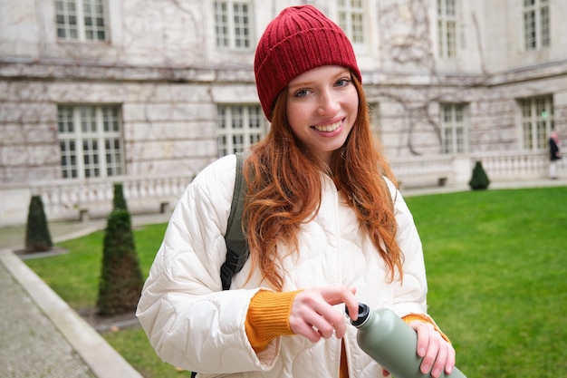 Gratis foto portret van een gelukkige meisjesreiziger die hete thee drinkt uit een thermoskan tijdens het wandelen en het verkennen van de stadstouri