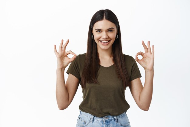 Portret van een gelukkige mooie vrouw die oke-tekens toont die lacht met OK-gebaren, keurt goed en houdt van reclame, prijst goede keuze die op een witte achtergrond staat