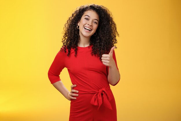 Portret van een gelukkige optimistische jonge vrouw met krullend haar in een rode jurk die vrolijk lacht, duimen opsteekt in goedkeuring en een leuk gebaar, opgetogen met een geweldig idee, een plan over een gele muur accepteert.