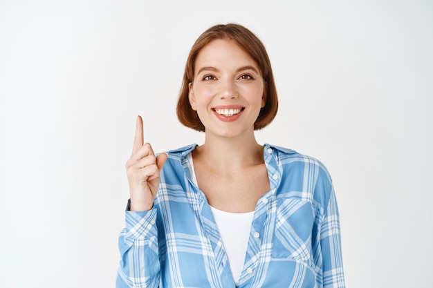 Portret van een gelukkige vrouw die lacht, vinger omhoog wijst, richting toont. Jong meisje dat speciale deal op witte muur aankondigt