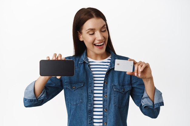 Portret van een gelukkige vrouwelijke klant, met creditcard, horizontaal smartphonescherm, tevreden kijkend naar korting, online winkelen, staande tegen een witte achtergrond.