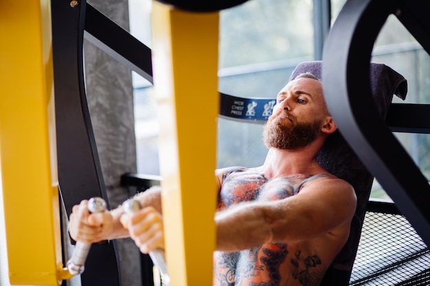 Portret van een getatoeëerde bebaarde man uit te werken op de persmachine van de borst in de sportschool in de buurt van venster