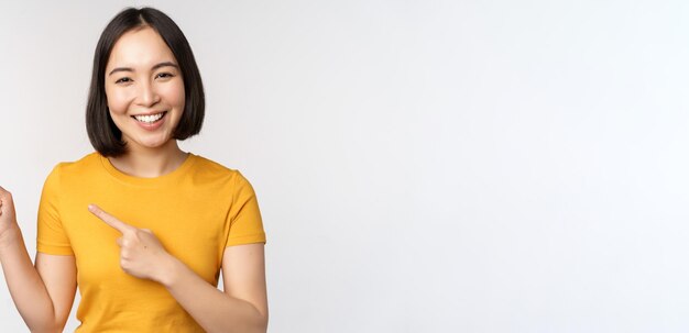 Portret van een glimlachend aziatisch brunette meisje in een geel t-shirt dat met de vingers naar links wijst en kopieerruimte p