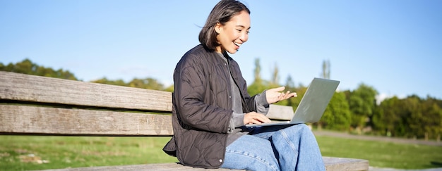 Gratis foto portret van een glimlachend aziatisch meisje dat op een bankje in het park zit en online met een vriend praat via laptop videochats