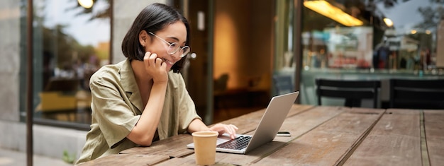 Portret van een glimlachend meisje in een bril die met een laptop op het terras zit, koffie drinkt en werkt