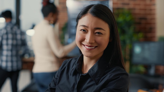 Portret van een glimlachende Aziatische softwareontwikkelaar die naar de camera kijkt in een ontwikkelingsbureau voor cloud computing. Zelfverzekerde programmeur poseren gelukkig naast collega's die werken in big data office.