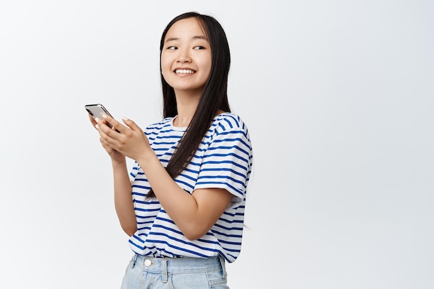 Portret van een glimlachende aziatische vrouw die een smartphone vasthoudt en er gelukkig uitziet met een applicatie op een mobiele telefoon die op een witte achtergrond staat