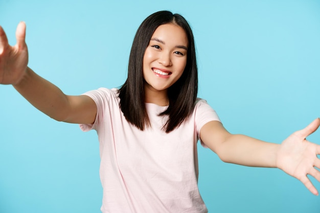 Portret van een glimlachende aziatische vrouw die naar een knuffel reikt, de handen vriendelijk spreidt, iets groots vasthoudt, over een blauwe achtergrond staat