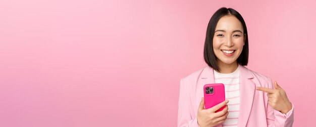 Portret van een glimlachende aziatische zakenvrouw die naar haar mobiele telefoon wijst en een app-app voor smartphones aanbeveelt op een mobiele telefoon die over een roze achtergrond staat