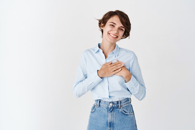 Portret van een glimlachende blanke vrouw die handen op het hart houdt en je bedankt, je dankbaar voelt, op een witte muur staat