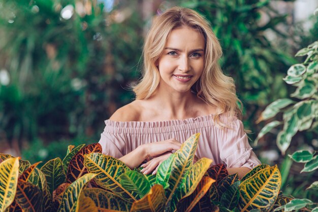 Portret van een glimlachende blonde jonge vrouw die zich achter de tuin croton bladeren bevindt