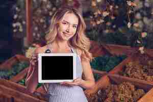 Gratis foto portret van een glimlachende blonde jonge vrouwelijke bloemist die digitale tablet met zwarte het schermvertoning toont