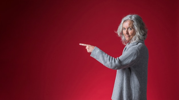 Portret van een glimlachende hogere vrouw die haar vinger richt tegen rode achtergrond