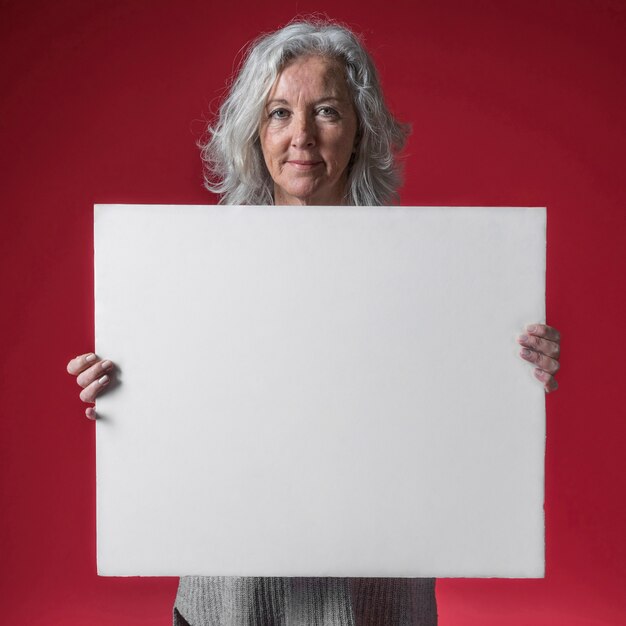 Portret van een glimlachende hogere vrouw die wit leeg aanplakbiljet toont tegen rode achtergrond