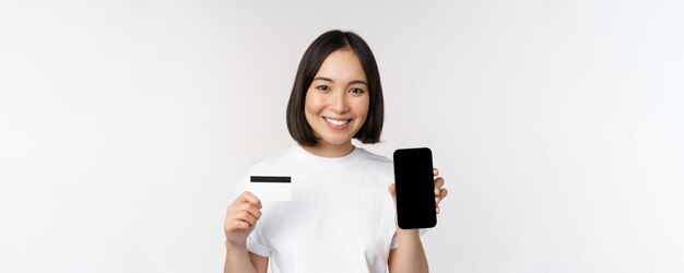 Portret van een glimlachende jonge aziatische vrouw die het scherm van de mobiele telefoon en een creditcard op een witte achtergrond toont