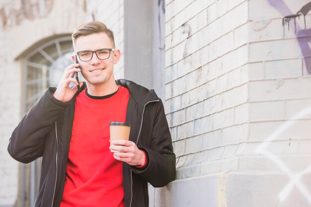 Portret van een glimlachende jonge mens die op mobiele telefoon spreekt die meeneemkoffie houdt